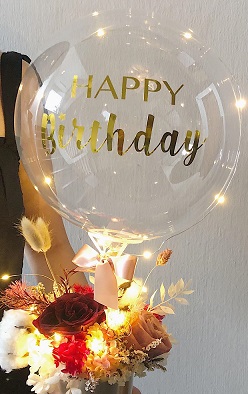 Happy birthday print on the transparent balloon with string lights and 12 flowers in a box